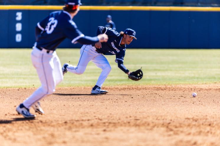 A Longwood baseball player in a navy uniform fields a ground ball, reaching low with his glove as a teammate moves nearby on the infield.