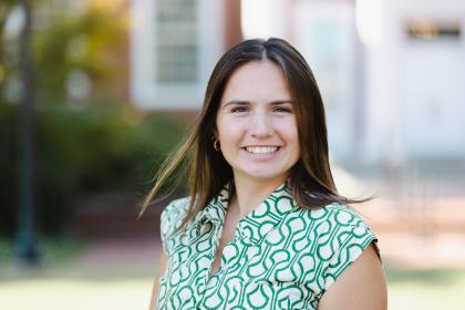 A smiling staff member stands outdoors in a close-up portrait on campus. She wears a green patterned blouse and small gold hoop earrings. Her long brown hair is lightly blown by the breeze. Campus buildings and greenery appear softly blurred in the background.