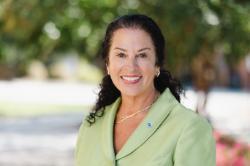 A woman with dark curly hair smiles outdoors, wearing a light green jacket and gold jewelry, with trees and flowers softly blurred in the background.