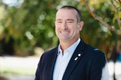 A man with closely cropped hair smiles outdoors, dressed in a navy blazer and light checkered shirt, with sunlight filtering through trees behind him.