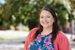 A woman smiles outdoors wearing a coral cardigan over a blue floral blouse, with soft green trees blurred in the background.