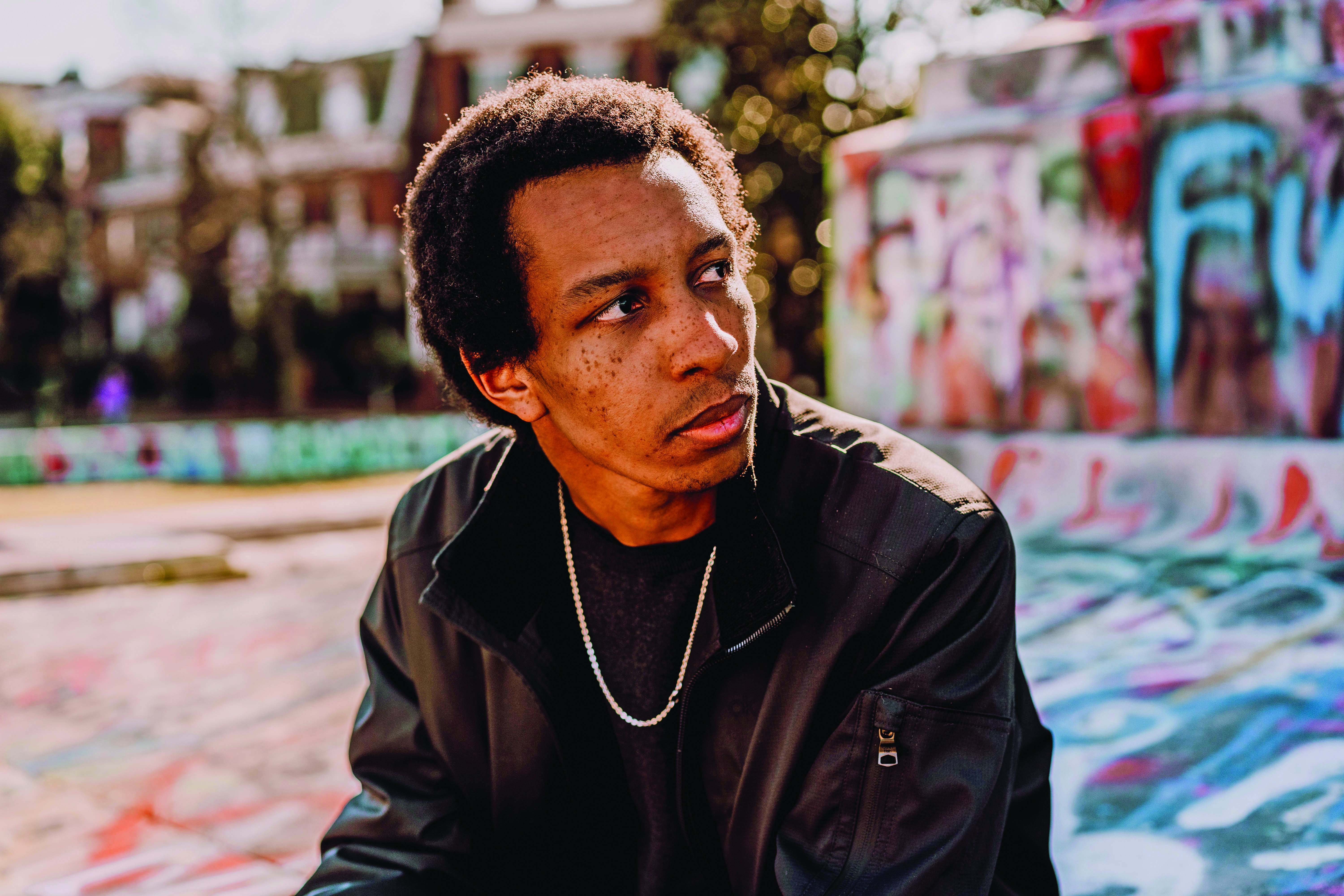 Joel Worford '18, credit: Maggie Graff/Ignited Creative 46 Man wearing a dark jacket and chain necklace sits outdoors near a graffiti-covered wall, looking off to the side with a thoughtful expression. Sunlight highlights his face, and blurred buildings and trees are visible in the background.