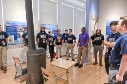 A museum guide in a purple polo shirt speaks to a group of Call Me MiSTER students gathered around a historic schoolroom exhibit, featuring vintage desks, a wood-burning stove, and informational panels about faculty and staff in segregated schools.