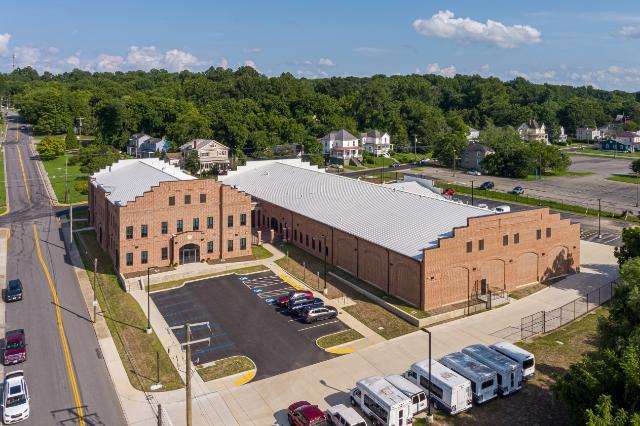 Aerial view of a large brick building with a white roof and stepped side design, surrounded by a parking lot with cars and RVs, residential houses, and green trees under a partly cloudy sky.