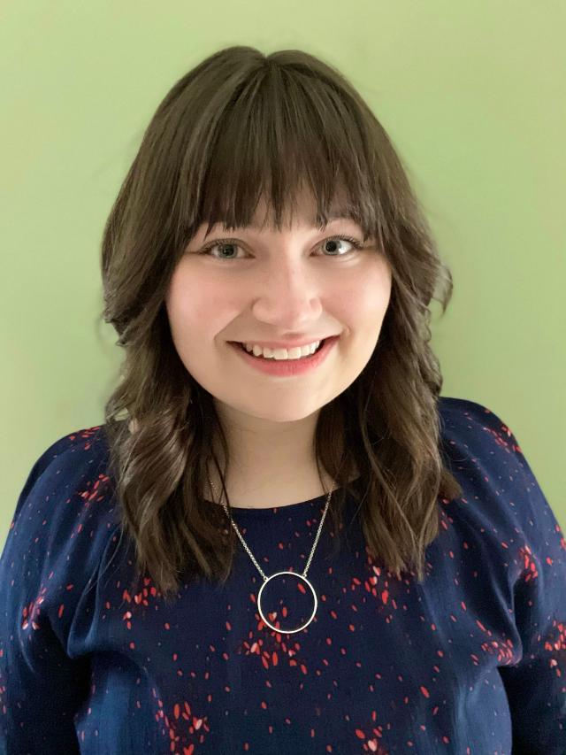Headshot of Megan Kruzinski the 2021-2022 recipient of the SNVC Scholarship award  - Woman with shoulder-length brown hair and bangs smiles in front of a light green wall. She wears a navy blouse with red speckles and a silver necklace with a circular pendant. Soft, even lighting highlights her face and gentle expression. The portrait has a warm, approachable, and professional feel.