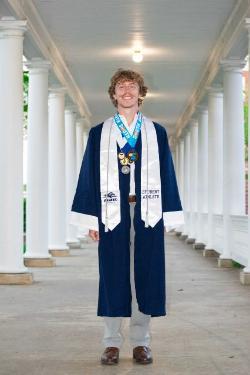 A graduate wearing a navy blue gown with white trim and stoles stands in a columned walkway. Medals hang around his neck, and he smiles at the camera under soft overhead lighting.
