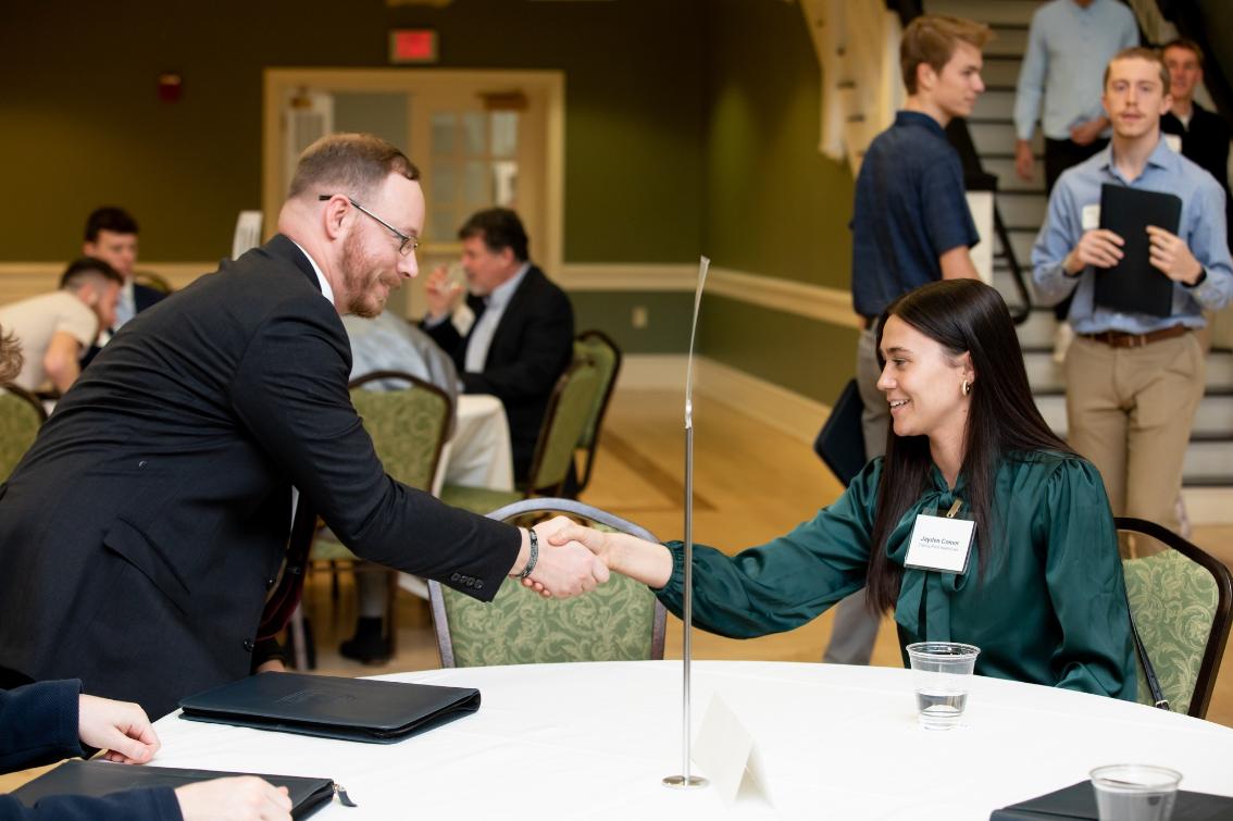Career Cafe 2 - A student seated at a table shakes hands with a professional during a networking event in a formal setting. Both are smiling and wearing name tags, while other attendees mingle and talk in the background.