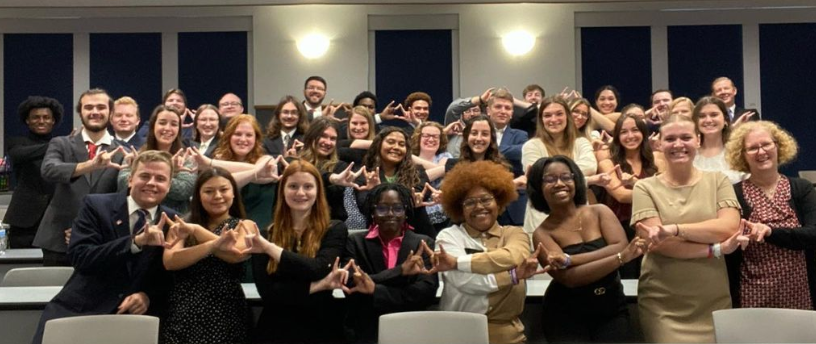Group photo of Kappa Nu brothers. - Large group of people posing together in a classroom. They smile and form a hand sign with fingers joined in front. Most wear business or semi-formal attire in coordinated rows. Bright lighting and large windows with dark blinds frame the background.