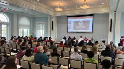 Education Immersion Day - image of an audience looking at a screen.  - A large, well-lit room with high ceilings and elegant lighting hosts an audience seated in rows facing a panel of six speakers beneath a screen displaying “Education for Equity: Panel Discussion.”