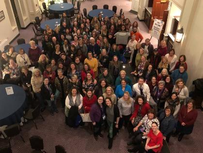 Longwood grad program represents at VAASL - A large group poses in a well-lit room with round tables and blue cloths.   Attendees of various ages wear name tags and lanyards.   The setting suggests a formal or semi-formal event or conference.   The background includes high ceilings and additional seating areas.
