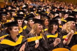 Graduate Commencement Ceremony - A group of graduates in black gowns and yellow stoles sit in rows.   They face forward with some clapping and smiling during the event.   The background shows an audience seated behind the graduates.   The setting reflects a formal and celebratory graduation ceremony.
