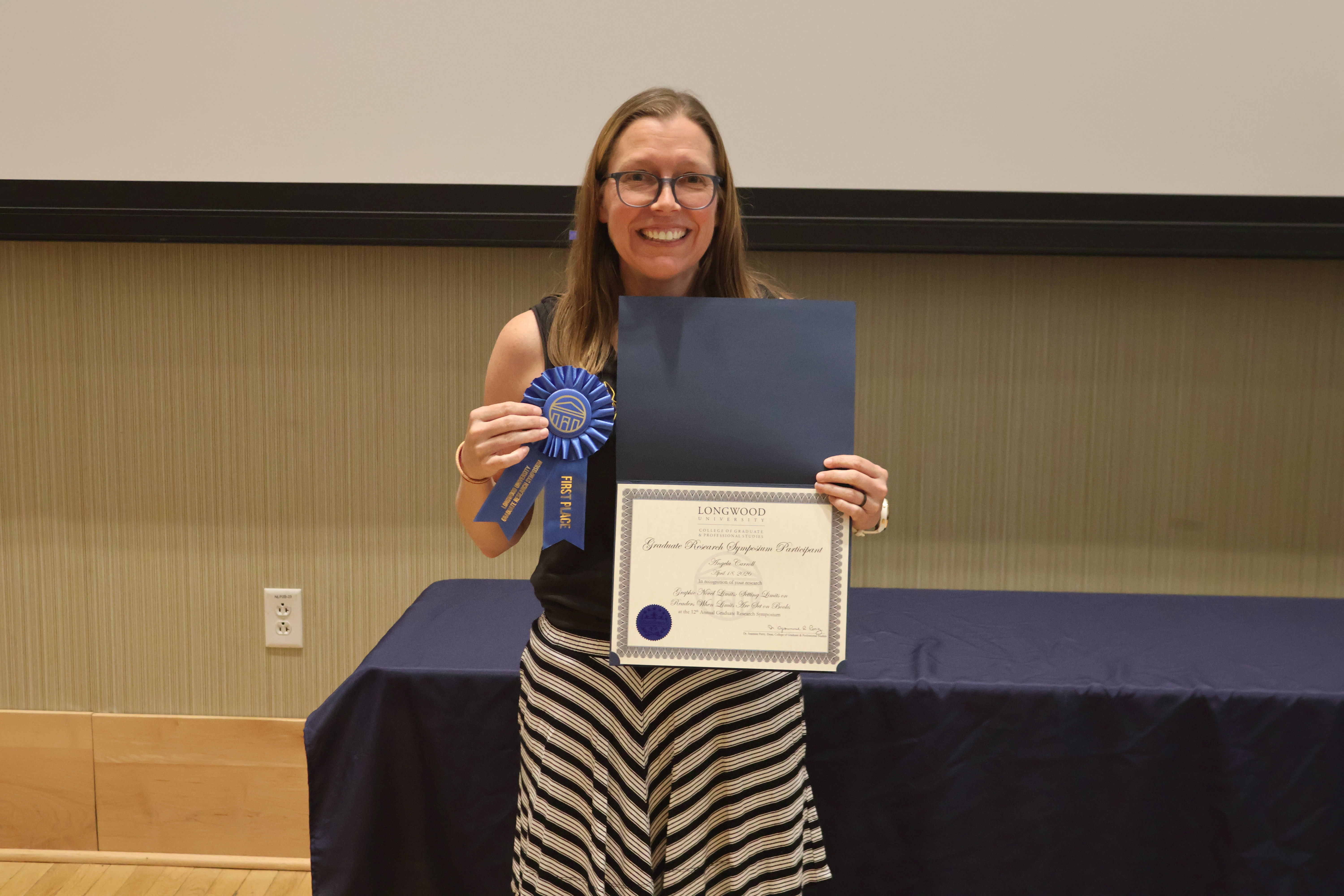- A Longwood graduate smiles while holding a certificate and first-place award ribbon.