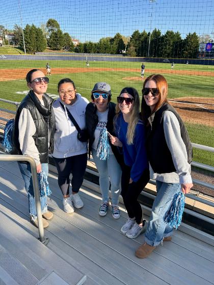 Baseball Night - Five people stand on bleachers at a baseball field holding pom-poms.   They wear casual clothes and sunglasses, showing team spirit.   The field, players, scoreboard, and trees are visible in the background.   The scene captures enthusiastic spectators enjoying the game.