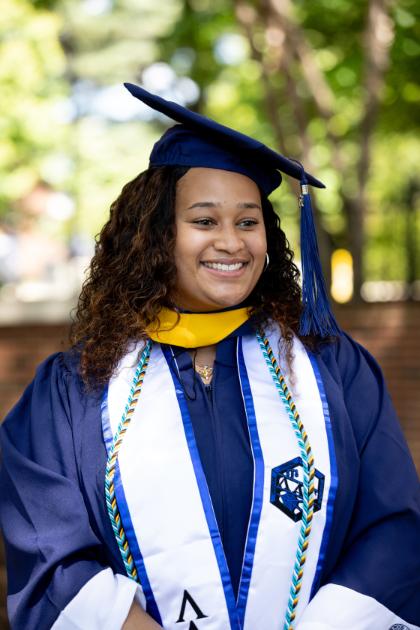 A student stands wearing academic regalia and smiles looking out of the frame.  - Graduate in a navy cap and gown smiles outdoors under shaded trees. She wears a yellow hood and a white stole with blue trim and embroidered symbols, along with honor cords. Soft sunlight filters through the leaves behind her, creating a warm, celebratory scene.