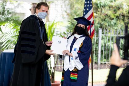 Class of 2020 graduate with President Reveley at the small personal graduation recognition ceremony - A graduate in a blue gown and white stole poses with a university official. Both wear masks while holding a diploma cover together during the ceremony. The American flag and lush greenery form the backdrop of the outdoor stage. The moment captures a proud graduation handshake under pandemic precau