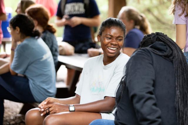 An honors student at the retreat laughs with her friends - Students sit together at picnic tables under a wooden shelter, enjoying a relaxed outdoor moment. A student in the foreground laughs warmly while talking with a friend. Others chat in small groups in the background, creating a lively, friendly atmosphere. The scene feels casual and welcoming, with natural light filtering through the trees.