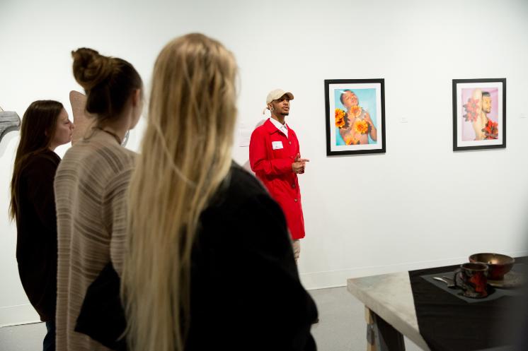 a student talks about their art work at research day - A student wearing a bright red coat presents their artwork to a small group in a gallery setting. Two framed portrait photographs featuring vibrant floral elements hang on the wall behind them. Attendees stand attentively, listening to the artist’s explanation. The scene conveys creativity and engagement during a campus research or art showcas