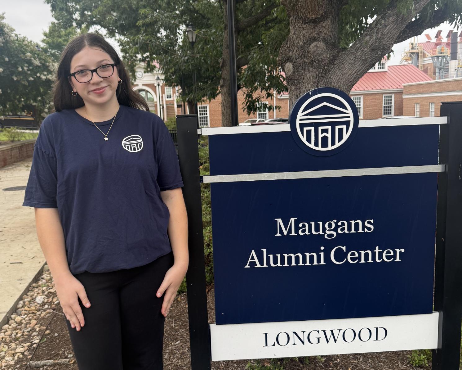Carly smiling to the side of a sign for the Maugans Alumni Center