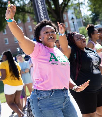 Longwood students at an event on campus - A student wearing a pink Alpha Kappa Alpha shirt dances joyfully outdoors with both arms raised. She smiles brightly in the sunlight, surrounded by other students enjoying the event. Another student beside her claps along, adding to the lively atmosphere. A Longwood University banner and campus buildings are visible in the background.