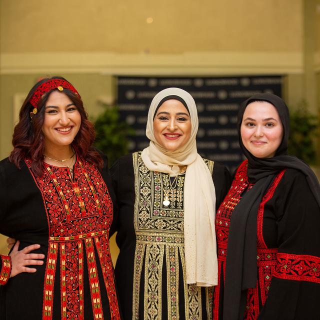 - Three women stand together smiling, dressed in traditional embroidered garments. Two wear red and black patterns, and one wears a cream hijab with gold and black embroidery. They pose closely in a warmly lit indoor space. A blurred backdrop with dark blue and white text stands behind them.