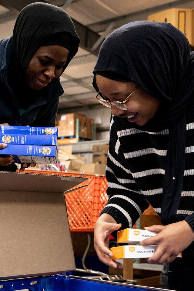 - Two students lean over a box while sorting packaged food items. Both wear headscarves and smile as they work together. One holds several boxes of macaroni and cheese over an open cardboard box. Shelves and supplies in a warehouse setting fill the background.
