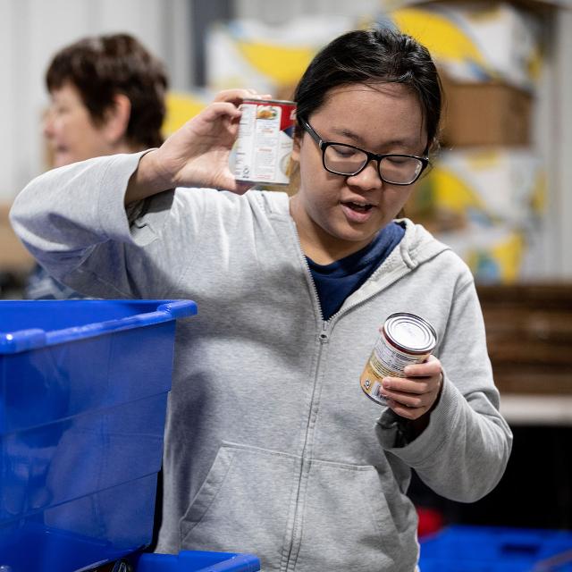 A student wearing glasses and a gray hoodie sorts canned goods. They hold one can up while placing another into a blue bin. Shelves and boxes fill the background of the warehouse space. The student appears focused and engaged in the service activity.