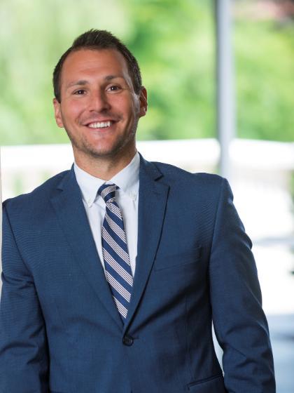 Smiling man in a blue suit and striped tie, standing in front of a blurred green and white background.