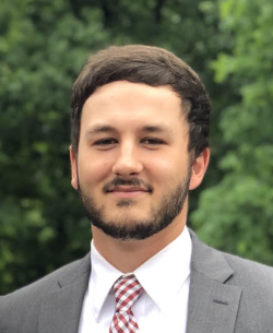 Headshot of Brennan Hoffer - A man in a gray suit jacket, white dress shirt, and red-and-white patterned tie smiles slightly while standing outdoors in front of a backdrop of green trees.