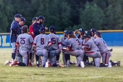 Brenna Hoffer in a huddle with baseball players. - A baseball team wearing gray pinstripe uniforms kneels in a huddle on the field while coaches stand around them, all listening intently during a meeting near the outfield fence.
