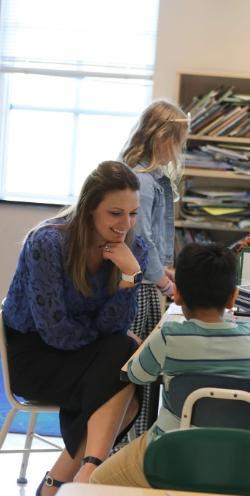 A teacher kneels beside a young student at a desk, smiling as they talk. She rests her chin on her hand while listening and offering guidance. Another child stands nearby in the classroom. Bookshelves and a bright window fill the background.