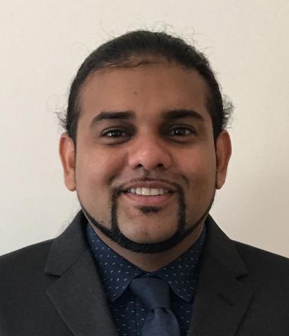 Headshot of Jason Kissoon - A man with dark hair pulled back smiles at the camera, wearing a dark suit jacket, a navy patterned shirt, and a matching tie against a plain light background.
