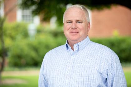 - A man with short light hair stands outdoors in soft, natural light. He wears a light blue checked button-down shirt and smiles gently at the camera. Green trees and shrubs create a blurred background behind him. A brick campus building is partially visible in the distance.