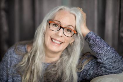 A woman with long gray hair and glasses with orange accents smiles warmly, leaning her head on her hand against a soft, dark background.
