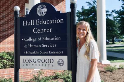 MaddieHommey stands next to the Hell Education Center sign.  - A smiling woman in a white dress stands beside the Hull Education Center sign on Longwood’s campus. Sunlight highlights her long blonde hair as she poses next to the black and white sign. Brick walls, columns, and greenery frame the background. The scene feels bright and welcoming outdoors.
