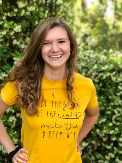 Sadie Hodges - A young woman with long, wavy brown hair smiles brightly while standing outdoors. She wears a yellow T-shirt with the text “See the good, be the light, make the difference.” Green foliage and small white flowers create a soft, natural background. Her hands rest on her hips, giving the portrait an upbeat and confident feel.