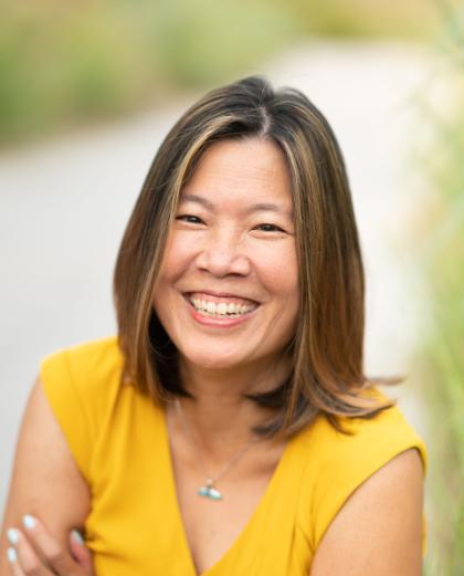 Smiling woman with shoulder-length hair wearing a yellow top and a necklace, standing outdoors with a blurred natural background.