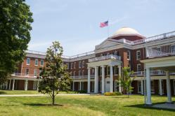 Rotunda - A stately red-brick campus building features white columns and a domed roof. An American flag waves above the central portico. Manicured lawns and young trees frame the front courtyard. Bright summer light highlights the building’s classical architecture.