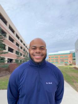 - A smiling man stands outdoors on a campus walkway. He wears a navy zip-up jacket with “Be Well” printed on the chest. Academic buildings and greenery are visible in the background. The portrait is framed at mid-chest with a soft, overcast sky behind him.