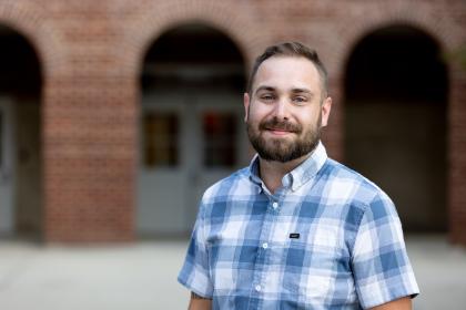 Dr. Jarod Vance - A bearded man stands outdoors in front of a brick building with arched walkways. He wears a blue-and-white plaid short-sleeve button-down shirt. The background is softly blurred, focusing attention on his face and upper body. The image appears to be a professional portrait taken on a college campus.