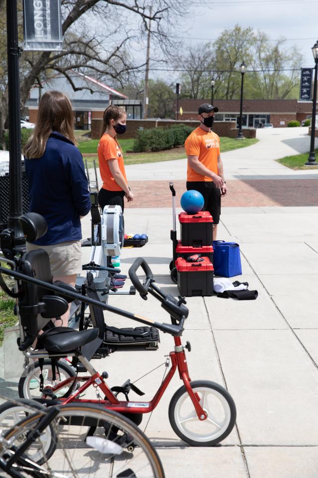 Sportable CTRS explains adaptive equipment. - A bearded man smiles at the camera during an outdoor portrait session. He wears a blue patterned button-down shirt with a light-colored tie. The background is softly blurred with green trees and campus elements. The image is a professional headshot with natural lighting.