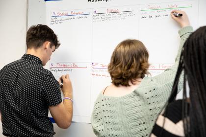 Students participating in interprofessional education events - Students write notes on a large whiteboard during a group activity. The board is divided into sections labeled Special Education, Nursing, and Social Work. Participants use markers to add ideas and assumptions under each category. The scene shows collaborative learning in an academic or career exploration setting.