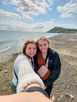 Two students take a selfie at the coast in Ireland. - Two students smile while taking a selfie on a rocky beach beside the ocean. The shoreline curves behind them with gentle waves and a distant hillside. One student extends an arm toward the camera, capturing the moment together. The scene shows a bright, breezy day along the coast.