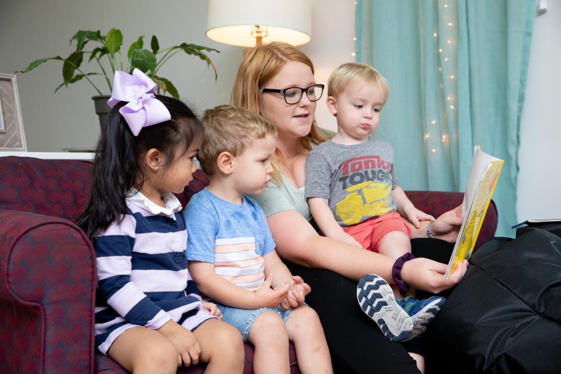 Teacher reading to three children at the Andy Taylor Center - A woman wearing glasses reads a children’s book while seated on a couch. Three young children sit close beside her and look toward the open book. A girl with a purple bow and two small boys listen during story time. A lamp, houseplant, and teal curtains create a cozy indoor setting.