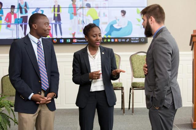 Three people in professional attire talk in a conference setting. They stand in front of a large screen showing colorful graphics of people working. One woman gestures as she speaks while the two men listen attentively. Chairs and presentation materials are visible in the background.
