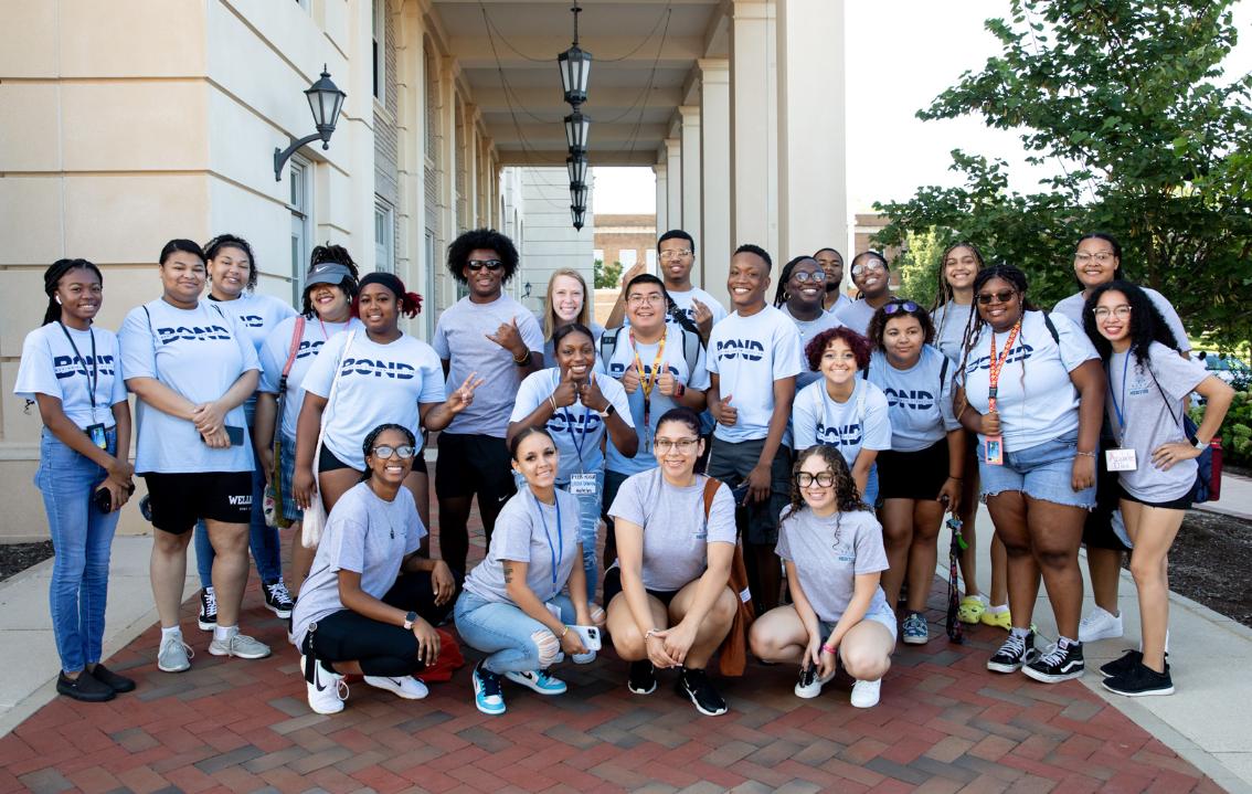 RISE mentors assisting with Move In Fall 2023 - A large group of students poses together outside a campus building. They wear matching “BOND” T-shirts and smile toward the camera. Some stand while others kneel in front, creating a group formation. Columns, hanging lights, and trees frame the background.