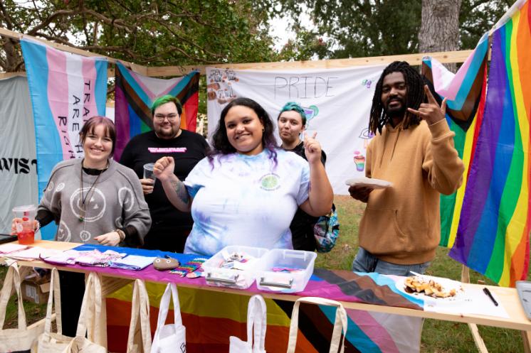 PRIDE booth at Oktoberfest - A group of students stands behind a table at a Pride-themed campus booth. Colorful flags and a banner reading “PRIDE” hang behind them. They smile and pose while displaying crafts and items on the table. Trees and an outdoor campus setting surround the scene.