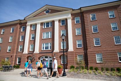 Sharp Hall - Students and families walk toward the entrance of a large brick residence hall. The building features tall white columns and rows of windows across several floors. People carry bags and belongings as they arrive during move-in. A sunny blue sky and landscaped walkway frame the campus scene.