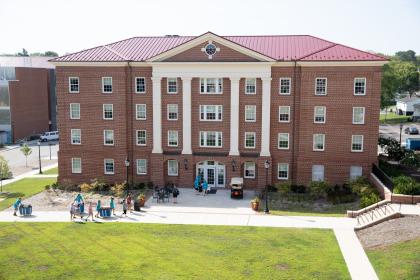 Register Hall - Large brick residence hall with white columns and a red metal roof. Students move belongings along the sidewalk in front of the building during move-in. Several people gather near the main entrance while others push carts with bins. Green lawn and walkways surround the building on a sunny day.