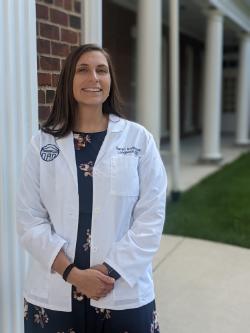 Photo of Sarah Anderson in a white lab coat. - A woman wearing a white lab coat stands smiling beside a brick column. The coat is embroidered with the name Sarah Anderson and Longwood. She stands outdoors near a walkway with white columns and green lawn. Her hands are clasped as she looks toward the camera.