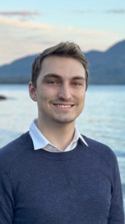 A smiling man with short brown hair stands outdoors by a calm lake. He wears a white collared shirt under a navy sweater. Mountains rise in the background under a soft, pastel sky. The portrait is tightly framed with the background gently blurred.